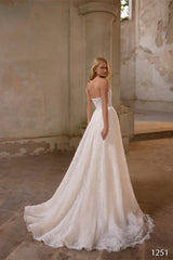 O femeie îmbrăcată în rochia de mireasă Eden Bride Rochie de mireasă V-1251, o rochie albă fără bretele, cu dantelă delicată și trenă lungă, stă în interior lângă coloane înalte, în lumina naturală, cu spatele la cameră și privind peste umăr. În colț apare inscripția „V-1251”. Rochii de mireasa Eden Bride