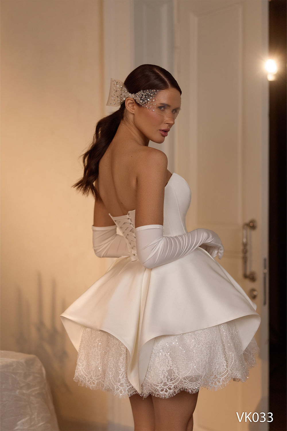 O femeie îmbrăcată în rochia de mireasă scurtă „Eden Bride” modelul VK-033, cu un top alb fără bretele, o fustă evazată și structurată peste dantelă, mănuși albe lungi și un accesoriu decorativ pentru păr, stă în interior, lângă o ușă, privind peste umăr. Rochii de mireasa Eden Bride