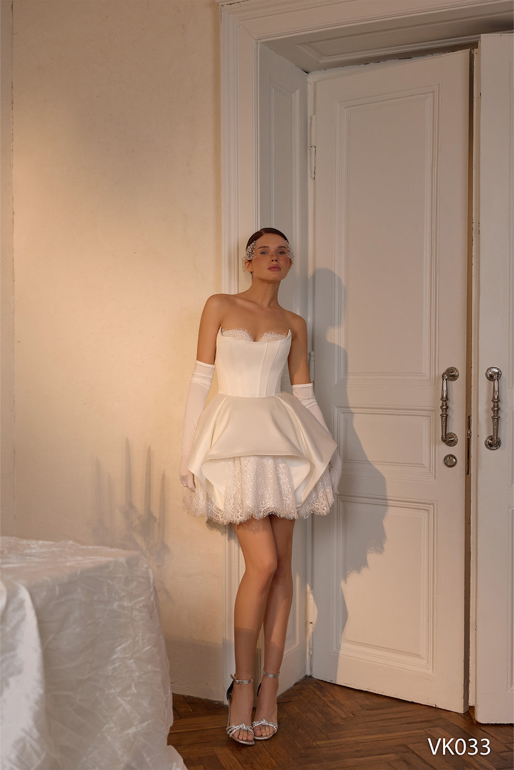 O femeie poartă rochia de mireasă scurtă Eden Bride VK-033, cu borduri delicate din dantelă, mănuși albe lungi și o coroniță împodobită cu pietre prețioase, în timp ce stă lângă ușile duble, înconjurată de o lumină difuză și un decor elegant, privind ușor în sus. Rochii de mireasa Eden Bride