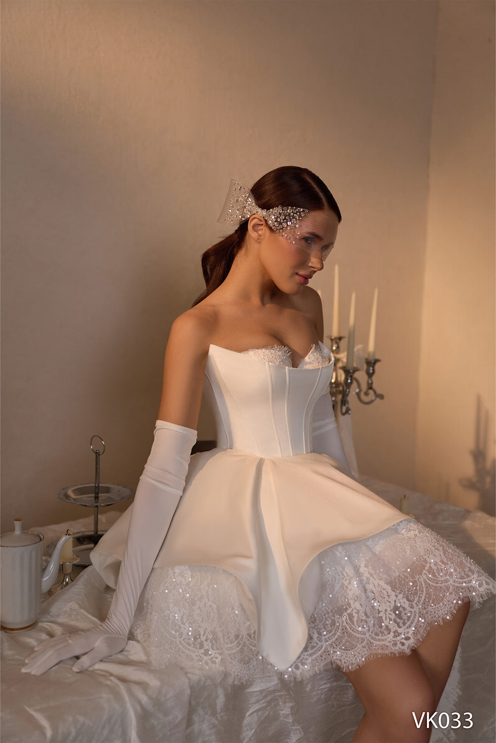 O femeie îmbrăcată în rochia de mireasă scurtă Eden Bride VK-033, împodobită cu detalii din dantelă și un accesoriu strălucitor pentru păr, stă lângă o masă pe care se află un ceainic și un candelabru, purtând mănuși lungi și privind în jos cu un aer gânditor. Rochii de mireasa Eden Bride