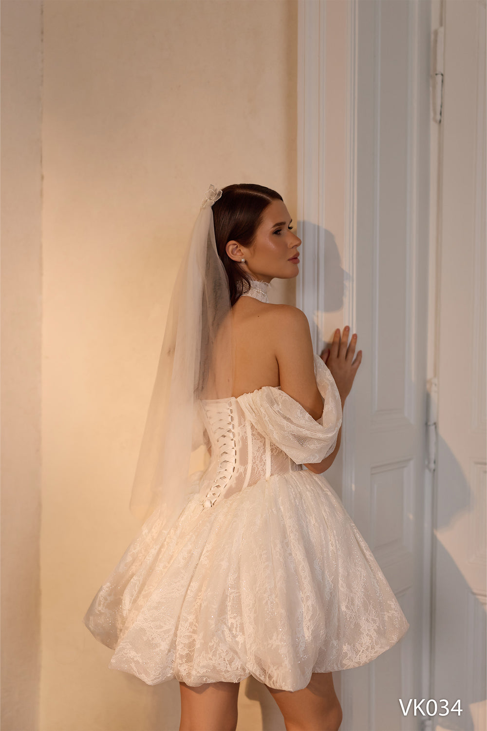 O femeie îmbrăcată în rochia de mireasă scurtă Eden Bride VK-034, cu dantelă și voal, stă cu profilul lângă o ușă cu panouri albe, atingând-o ușor, în timp ce o lumină caldă iluminează discret scena. Rochii de mireasa Eden Bride