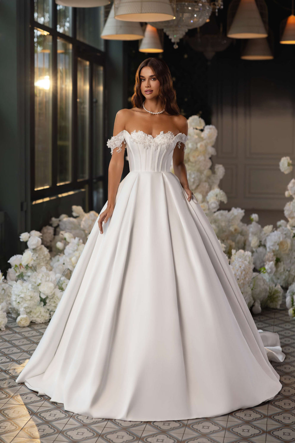 O femeie în eleganta Rochie de mireasa Eliana by Eden Bride, cu un corset din dantelă și un design fără umeri, stă în interior printre flori albe și lumini moi, ferestre mari și lumini suspendate în spatele ei. Rochii de mireasa Eden Bride