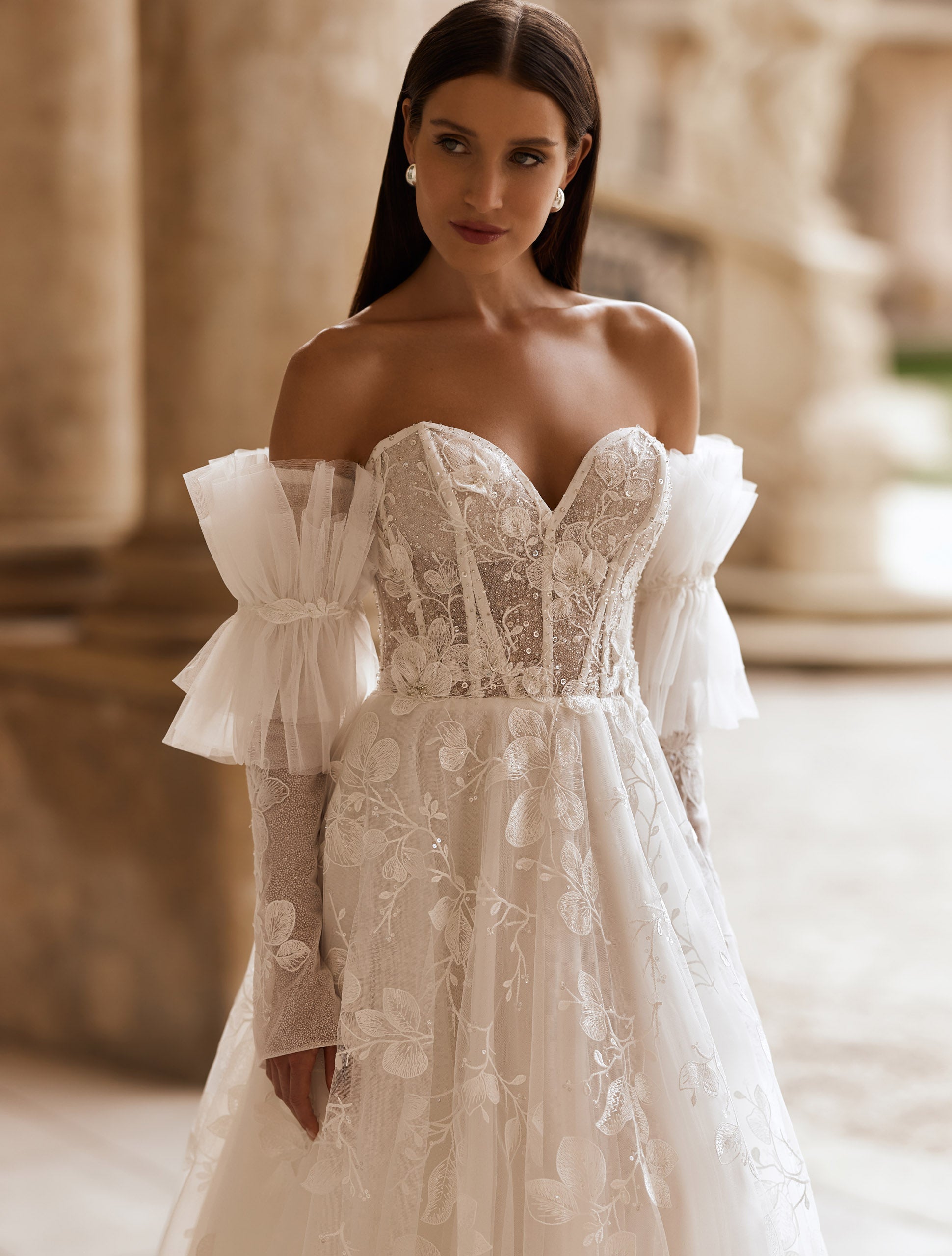 O femeie îmbrăcată în rochia de mireasă Eden Bride Rochie de mireasă J-1118 — o rochie de mireasă sofisticată din dantelă, cu umerii goi, cu un corset în formă de inimă, broderii florale transparente și mâneci spectaculoase din tul — stă în aer liber, în fața unei clădiri clasice din piatră. Rochii de mireasa Eden Bride