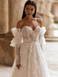 O femeie îmbrăcată în rochia de mireasă Eden Bride Rochie de mireasă J-1118 — o rochie de mireasă sofisticată din dantelă, cu umerii goi, cu un corset în formă de inimă, broderii florale transparente și mâneci spectaculoase din tul — stă în aer liber, în fața unei clădiri clasice din piatră. Rochii de mireasa Eden Bride