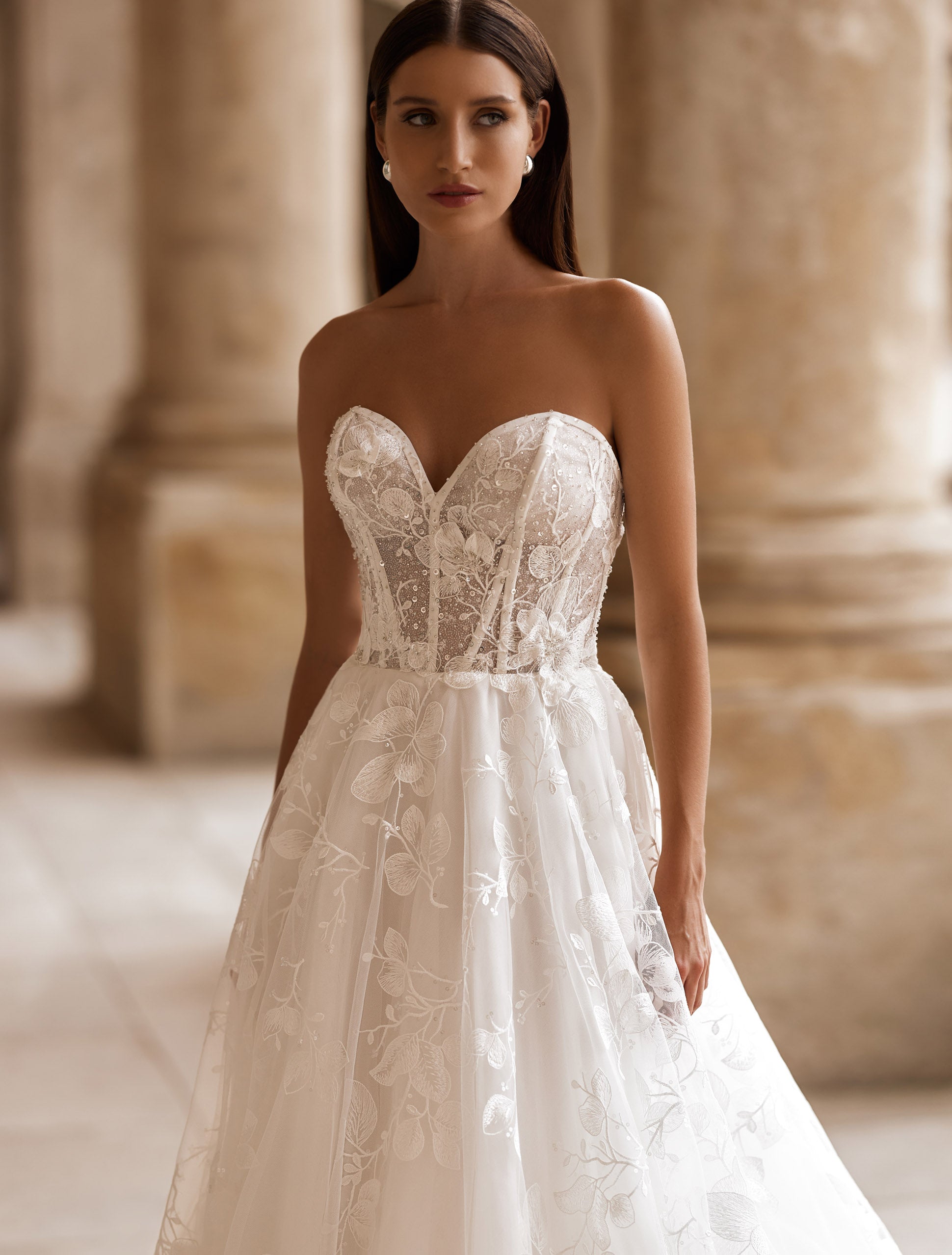 O femeie îmbrăcată în rochia de mireasă „Eden Bride Rochie de mireasă J-1118”, o rochie albă de mireasă fără bretele, cu croi în A și broderie din dantelă, stă într-un coridor din piatră cu coloane, privind ușor în lateral. Rochii de mireasa Eden Bride