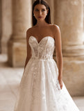 O femeie îmbrăcată în rochia de mireasă „Eden Bride Rochie de mireasă J-1118”, o rochie albă de mireasă fără bretele, cu croi în A și broderie din dantelă, stă într-un coridor din piatră cu coloane, privind ușor în lateral. Rochii de mireasa Eden Bride