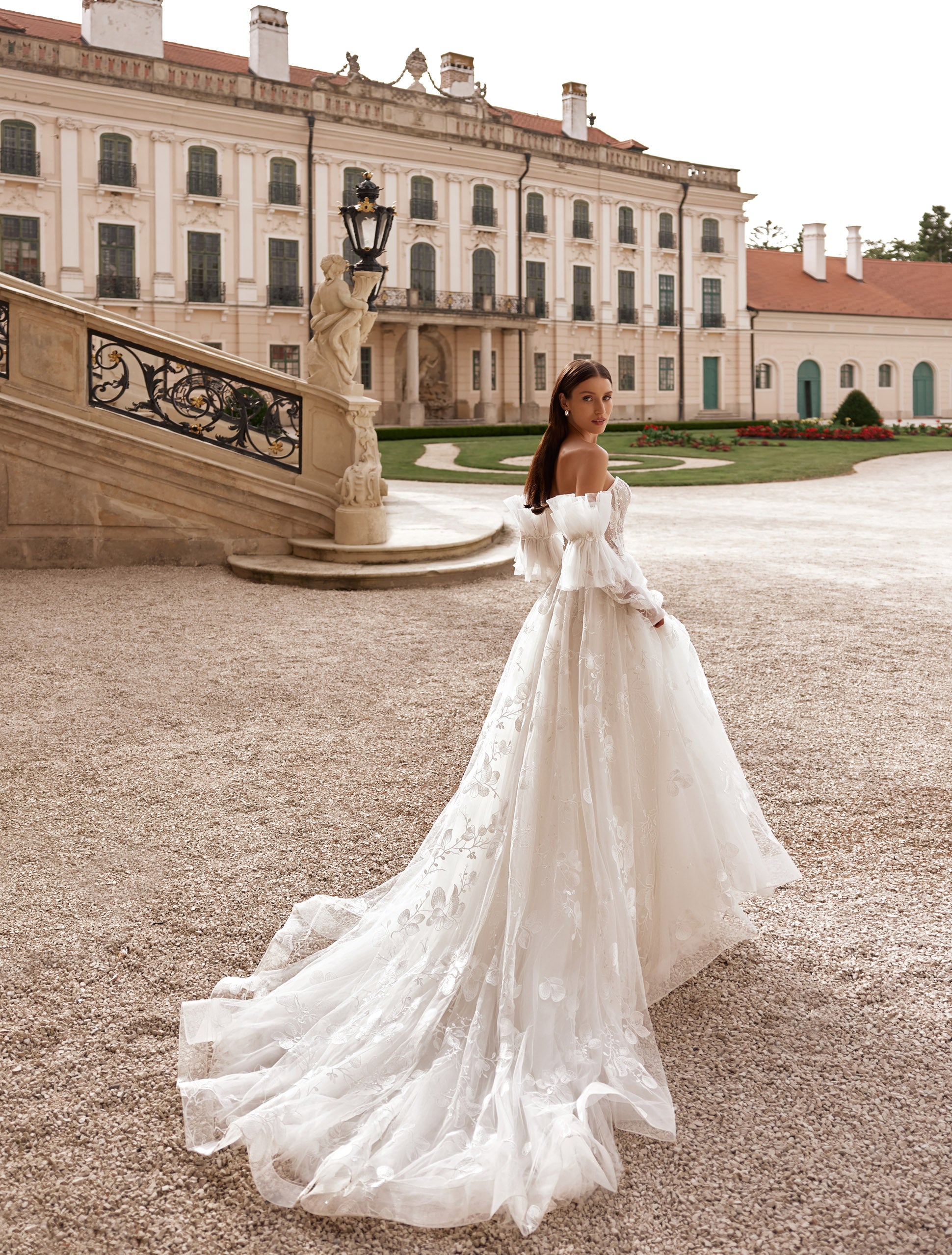 O femeie îmbrăcată în rochia de mireasă J-1118 de la Eden Bride, o rochie de mireasă elegantă în linie A, cu corset în formă de inimă, stă în aer liber lângă o clădire istorică impunătoare, cu o arhitectură bogat ornamentată și o scară de piatră. Rochii de mireasa Eden Bride
