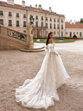 O femeie îmbrăcată în rochia de mireasă J-1118 de la Eden Bride, o rochie de mireasă elegantă în linie A, cu corset în formă de inimă, stă în aer liber lângă o clădire istorică impunătoare, cu o arhitectură bogat ornamentată și o scară de piatră. Rochii de mireasa Eden Bride