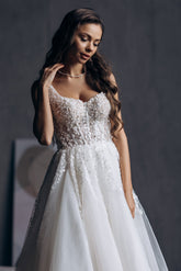 O femeie în Eden Bride Rochie de mireasa Bella, o linie A albă fără mâneci cu dantelă florală și o fustă plină de tul, stă pe un fundal întunecat, privind în jos gânditor și atingându-și colierul. rochii de mireasa Eden Bride