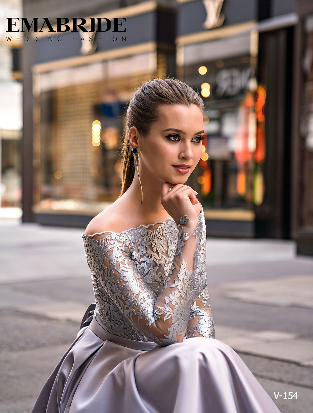 O femeie într-un Eden Bride ROCHIE DE SEARA argintiu V-154 cu dantelă florală stă în aer liber, zâmbind gânditoare. În fundal apar vitrine elegante și textul EMABRIDE WEDDING FASHION și V-154. Rochii de mireasa Eden Bride