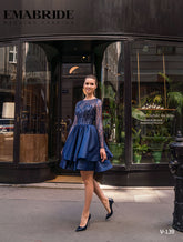 O femeie într-un ROCHIE DE SEARA V-139 scurt, albastru marin, cu mânecă lungă, de la Eden Bride și tocuri zâmbește pe un trotuar de oraș în fața unei vitrine elegante. Ideal pentru cununie civila sau evenimente elegante. Logo-ul Eden Bride apare în partea de sus. Rochii de mireasa Eden Bride