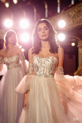 Două femei poartă ROCHIE DE SEARA EN V-534 de la Eden Bride, rochii albe elegante, fără umeri, cu broderii argintii, stând într-un decor ornamentat, luminat delicat. Una dintre ele privește în față; cealaltă apare ușor estompată în fundal. Rochii de mireasa Eden Bride