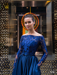 O femeie în Eden Bride ROCHIE DE SEARA V-154, o rochie de seară din dantelă albastru marin cu mâneci lungi, stă încrezătoare cu mâinile în buzunare în fața unui perete cu model elegant și a unei vitrine decorative moderne. Rochii de mireasa Eden Bride