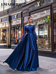 O femeie îmbrăcată în rochia Eden Bride ROCHIE DE SEARA V-154, o rochie navy lungă cu mâneci din dantelă și decolteu bateau, pozează elegant pe trotuarul unui oraș, în fața unor vitrine de lux, în timp ce rochia se desfășoară grațios. Rochii de mireasa Eden Bride