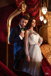 Un bărbat într-un smoching negru și o femeie purtând Eden Bride ROCHIE DE SEARA EN V-541 stau împreună într-un balcon de teatru ornat, roșu și auriu, cu iluminare elegantă și draperii bogate. Rochii de mireasa Eden Bride