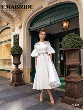 O femeie în eleganta rochie albă Eden Bride ROCHIE DE SEARA V-143 se află în fața unei clădiri elegante cu uși duble verzi, flancată de două tufe mari cu ghivece. Rochii de mireasa Eden Bride