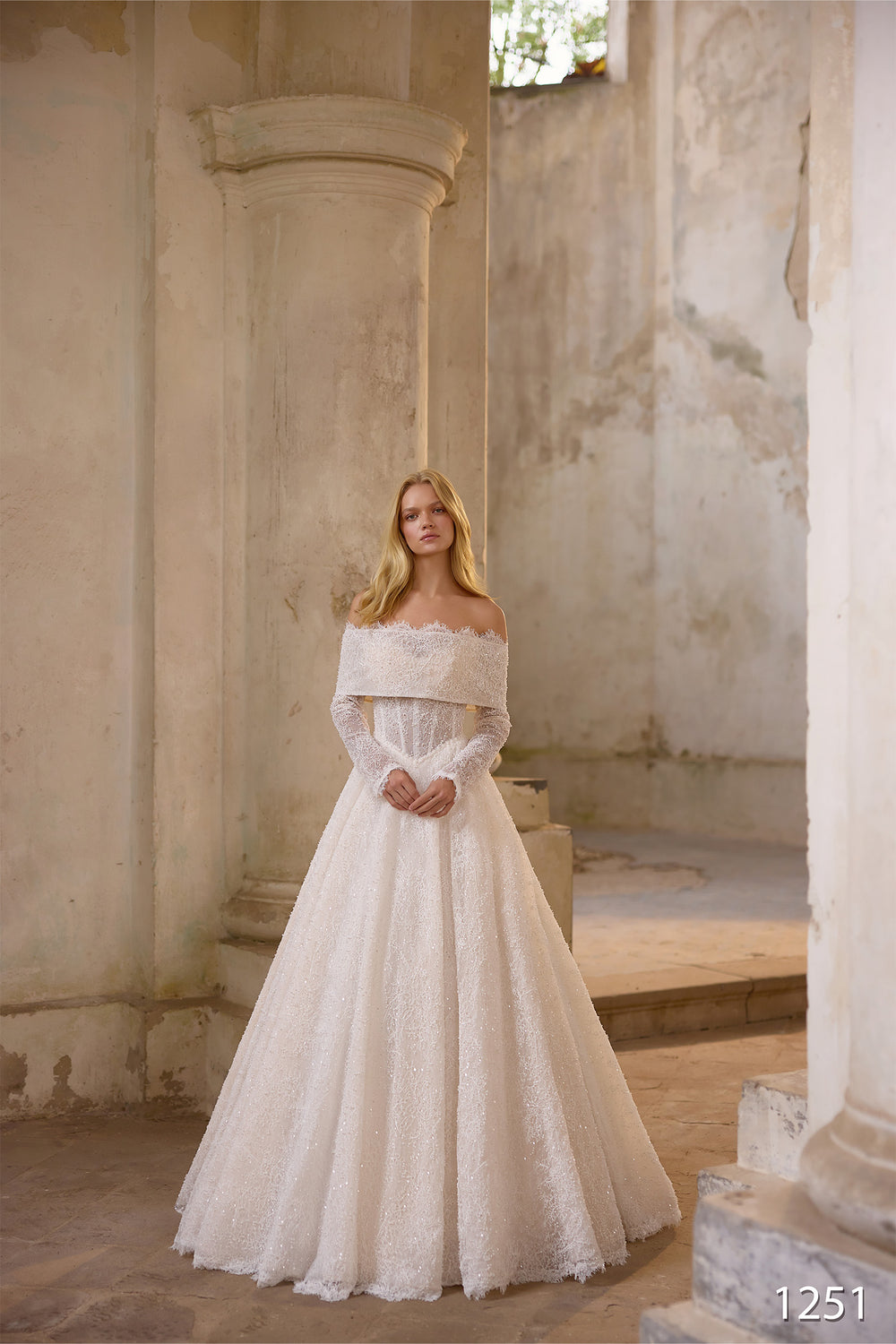 O femeie îmbrăcată într-o rochie de mireasă elegantă Eden Bride Rochie de mireasă V-1251, cu umerii descoperiți, mâneci lungi detașabile și dantelă fină, stă într-o clădire rustică din piatră, scăldată într-o lumină naturală blândă. Numărul 1251 apare în colțul din dreapta jos. Rochii de mireasa Eden Bride