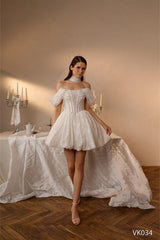 O femeie poartă rochia de mireasă scurtă VK-034 de la Eden Bride, cu corset din dantelă și fustă evazată, voal și pantofi cu toc. În fundal se văd lumânări aprinse și fețe de masă albe pe o masă decorată elegant. Rochii de mireasa Eden Bride