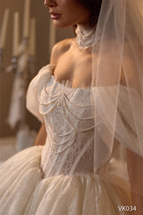 O mireasă poartă rochia de mireasă scurtă Eden Bride VK-034 — o rochie elegantă din dantelă, cu umerii descoperiți, cu accente de perle și un colier strâns pe gât din material transparent, asortată cu un voal. Lumina difuză a lumânărilor din fundal pune în valoare această rochie rafinată pentru căsătoria civilă. Rochii de mireasa Eden Bride