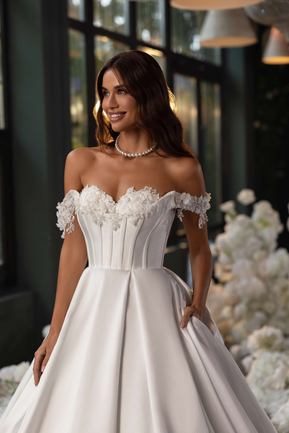 O femeie poartă Rochie de mireasa Eliana by Eden Bride, cu un corset dantelă off-the-shoulder și fustă prințesă, în interior cu un colier de perle și flori albe, în lumină naturală blândă. Rochii de mireasa Eden Bride