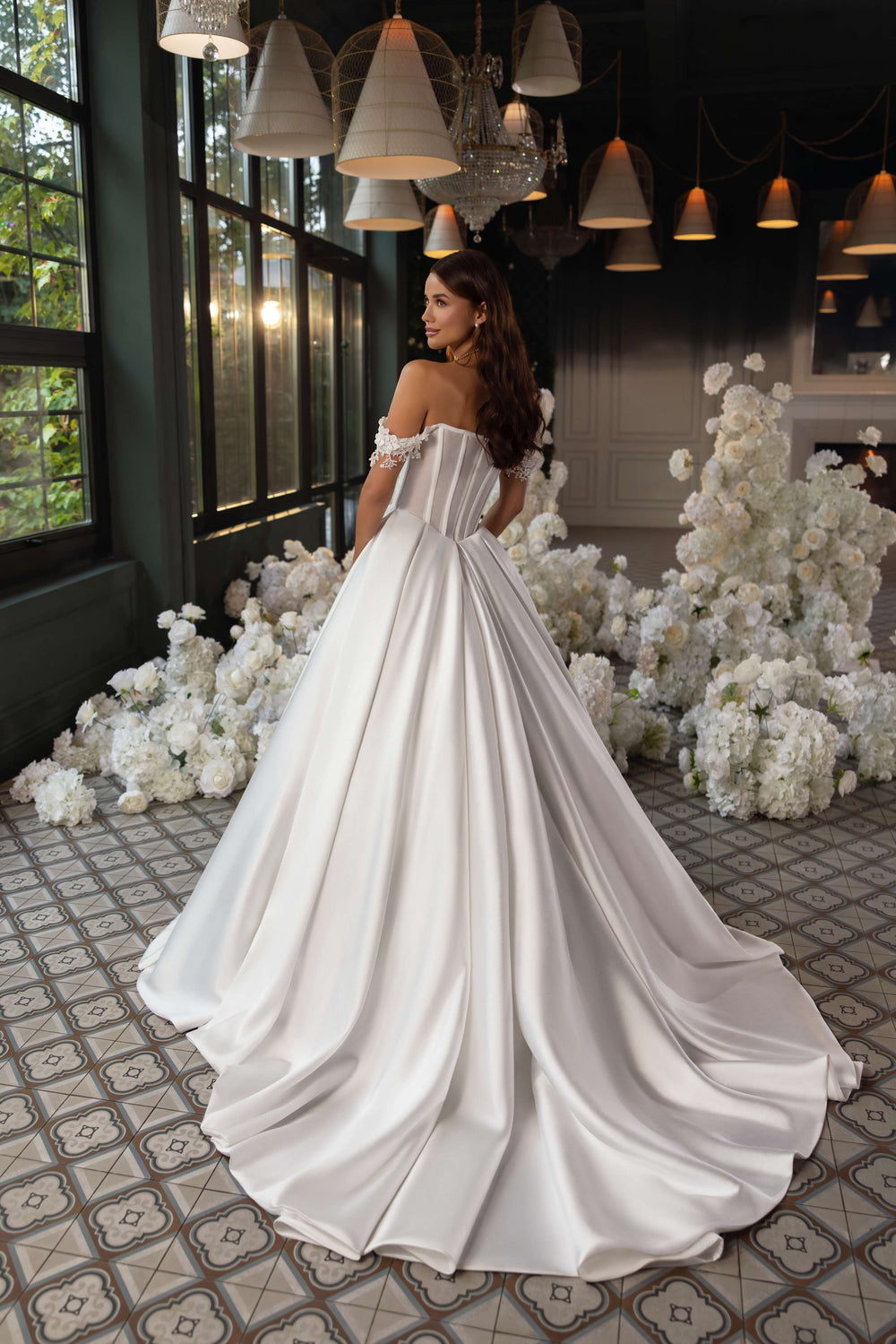 O femeie în elegantul Rochie de mireasa Eliana by Eden Bride, cu corset dantelă și fustă prințesă cu trenă fluidă, stă pe gresie cu model înconjurată de flori albe și ferestre mari într-o încăpere luminată ușor. Rochii de mireasa Eden Bride