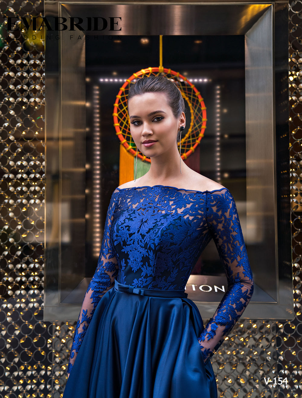 O femeie în Eden Bride ROCHIE DE SEARA V-154, o rochie de seară din dantelă albastru marin cu mâneci lungi, stă încrezătoare cu mâinile în buzunare în fața unui perete cu model elegant și a unei vitrine decorative moderne. Rochii de mireasa Eden Bride