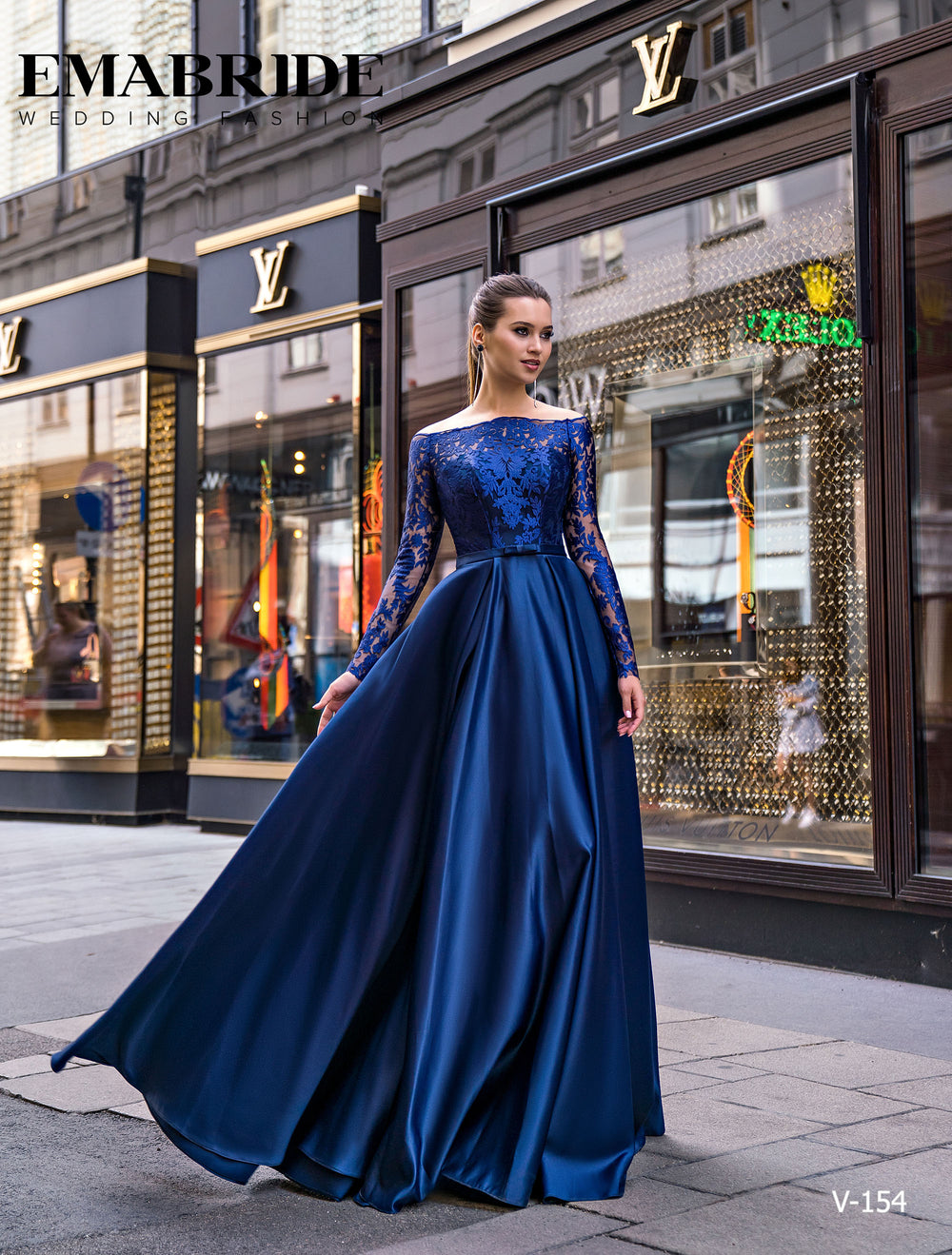 O femeie îmbrăcată în rochia Eden Bride ROCHIE DE SEARA V-154, o rochie navy lungă cu mâneci din dantelă și decolteu bateau, pozează elegant pe trotuarul unui oraș, în fața unor vitrine de lux, în timp ce rochia se desfășoară grațios. Rochii de mireasa Eden Bride