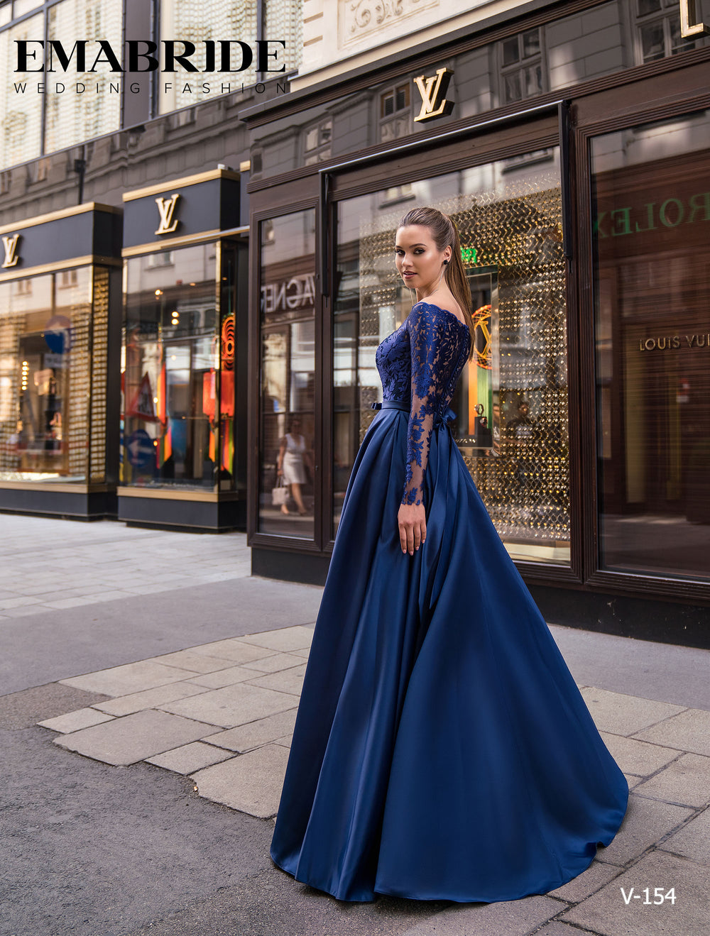 O femeie într-un ROCHIE DE SEARA V-154 albastru lung cu mâneci din dantelă de la Eden Bride stă pe o stradă a orașului în fața unui butic de lux - o alegere elegantă ca rochie de seara mama miresei. Rochii de mireasa Eden Bride